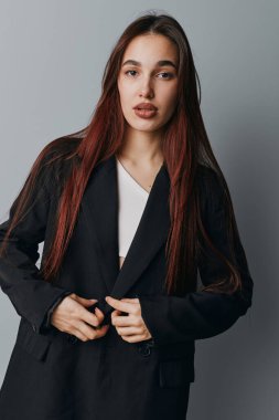 Gri arka planda duran siyah ceketli ve beyaz gömlekli uzun düz saçlı genç kadın portresi. Makyajı az olan kadın model sakin ve kendinden emin bir şekilde kameraya bakıyor..