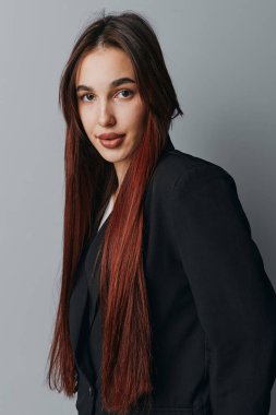 Gri arka planda duran uzun kahverengi saçlı, siyah ceketli genç bir kadının portresi. Makyajsız ve sakin yüzlü bir kadın model stüdyoda fotoğraf çekimi için poz veriyor..
