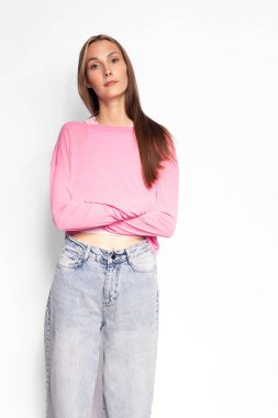 Pembe kazaklı ve kot pantolonlu kendine güvenen genç bir kadın beyaz arka planda çapraz kollarla poz veriyor. Sıradan moda, rahat tarz ve stüdyoda doğal güzellik kavramı..