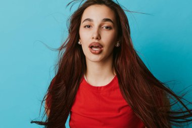 Genç bir kadının portresi, dalgalı kahverengi saçlı ve kırmızı başlıklı, mavi arka planda. Açık ağızlı ifade, doğal ten rengi, günlük kıyafetler, yaşam tarzı ve moda konsepti için dinamik saç..