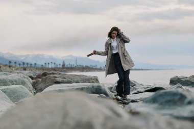 Uzun bir palto giyen genç bir kadın sakin deniz kıyısında büyük kayalarda dikkatle yürür. Bulutlu gökyüzü ve uzak dağlar kıyıya yakın sakin bir açık hava ortamı yaratır..