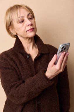 Kahverengi ceketli orta yaşlı kadın akıllı telefonu tutuyor ve sakin bir ifadeyle ekrana bakıyor. Bej renkli stüdyo. Dijital iletişim ve kıdemli yaşam tarzı teması.