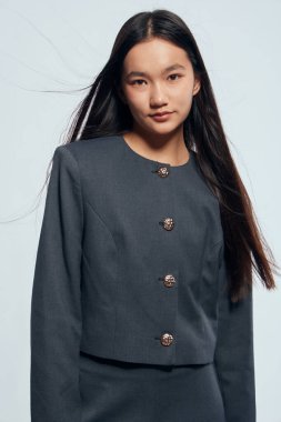 Dekoratif düğmeleri olan koyu renk takım elbiseli, sade bir arka planın önünde duran kendine güvenen genç bir kadın. Doğal makyajlı ve düz saçlı çağdaş tarz..