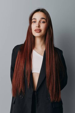 Siyah ceketli, beyaz bluzlu, kendine güvenen genç bir kadının portresi. Stüdyoda poz veriyor. Gri arkaplan, doğal makyaj ve günlük ifade.