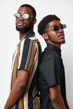 Güneş gözlüğü ve günlük kıyafetlerle esmer tenli iki siyah adamın portresi. Sırt sırta dururlar ve beyaz arka planı olan bir stüdyoda güven ve tarz gösterirler..