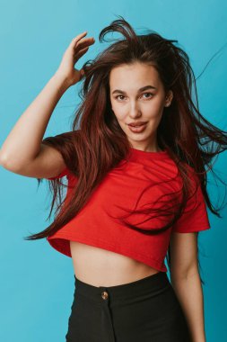 Kırmızı gömlekli, siyah pantolonlu, dalgalı saçlı, mavi arka planda duran ve kameraya gülümseyen ve el kaldıran bir kadın model..