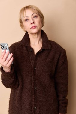 Kahverengi kazak giyen orta yaşlı ciddi bir kadın elinde bej arka planda duran akıllı telefonu tutuyor. Teknoloji, iletişim ve düşünceli yetişkin kavramı..