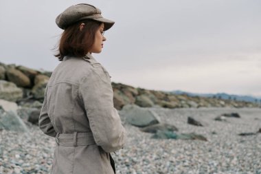 Bej ceketli ve şapkalı bir kadının kayalık bir kıyı şeridinde tek başına durup ufka baktığı görüntüler. Bulutlu gökyüzü ve soluk renkler yalnızlığı ve tefekküre vurgu yapar.