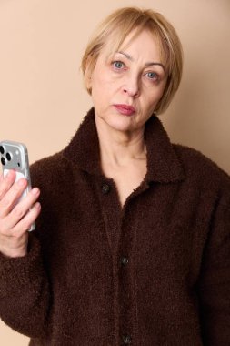 Kahverengi kazak giyen ciddi olgun bir kadın bej arka planda akıllı telefon tutuyor. İletişim, teknoloji kullanımı, duygular, orta yaş ve modern yaşam tarzı kavramı.