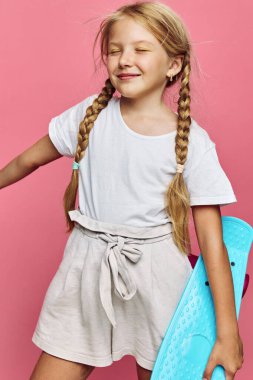 Beyaz tişörtlü, bej şortlu, iki örgülü gülümseyen kız pembe duvara karşı poz verirken mavi kaykay tutuyor. Gözler kapalı ve rahat. Gençlik, spor ve kaygısız anlar..