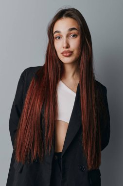 Siyah ceketli, beyaz bluzlu, düz kızıl saçlı genç bir kadın gri arka planda duruyor. Sıradan pozu olan şık bir bayan model ve minimal stüdyo ışıklandırması.