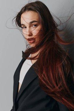 Kızıl kahverengi saçlı, soluk soluklu, resmi siyah ceketli ve beyaz gömlekli bir kadın model. Kapalı alanda kendine güvenen ifadesiyle kameraya bakıyor..