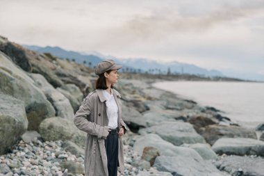 Örülmüş hırka ve kep giyen genç bir kadın kayalık sahilde okyanusa bakıyor. Bulutlu gökyüzü ve uzak dağlar doğal ortam ortamında barışçıl ve yansıtıcı bir hava oluşturur..