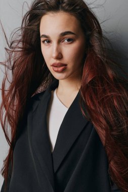 Uçan kızıl saçlı, ince makyajlı, siyah giysili, beyaz gömlekli, düz gri duvarın önünde duran genç bir kadın. Yumuşak doğal ışıkla yakın plan portresi.