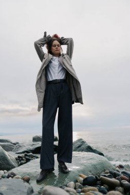 Sıcak paltolu ve siyah pantolonlu kadın bulutlu gökyüzünün altında okyanus kenarında taşların üzerinde poz veriyor. Sakin atmosfer, doğal manzara, moda kıyafeti ve rahat duruş açık havada çekildi..