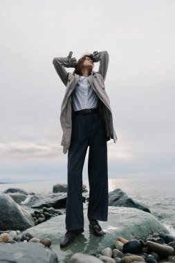 Uzun paltolu ve siyah pantolonlu şık bir kadın deniz kenarındaki kayalıklarda duruyor. Yukarı bakıyor, elleri başının üstünde, bulutlu gökyüzü ve sakin suyla çevrili. Açık hava tarzı ve havası.