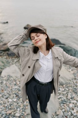 Bej ceket ve şapka giyen gülümseyen kadın çakıl taşı plajında gözleri kapalı duruyor. Sakin, doğa bağlantısı, özgürlük ve dışarıda suyun yanında huzurlu bir an..