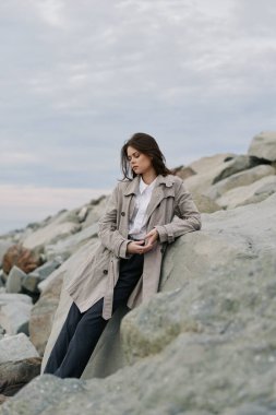 Trençkotlu ve bol pantolonlu kadın kayalık kıyı şeridine yaslanıyor. Bulutlu hava ve doğal manzara sakin ve içe dönük bir atmosfer yaratır. Tesadüfi zarafet ve doğa kavramı.