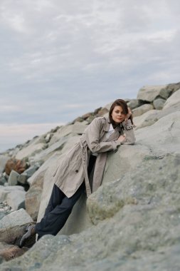 Sıcak palto giyen genç bir kadın bulutlu bir gökyüzünün altında okyanus kıyısındaki büyük kayalara yaslanıyor. Gündelik sonbahar modası, doğa bağlantısı, düşünceli ruh hali ve açık hava rahatlama kavramı..