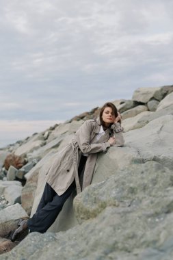 Rahat bir kazak ve siyah pantolon giyen genç bir kadın açık havada büyük kayalara yaslanmış pozlar veriyor. Bulutlu gökyüzü ve doğal kayalık ortam rahatlatıcı ve düşünceli bir atmosfer yaratır..