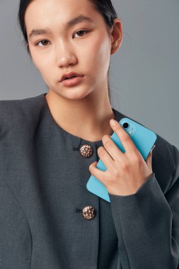 Sakin yüzlü yetişkin bir kadın göğsüne yakın turkuaz bir cep telefonu tutuyor. Kapalı alanda düğme detaylarıyla resmi kıyafetler giyiyor..