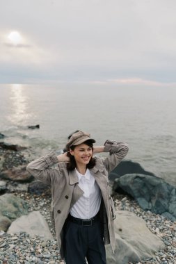 Şık palto ve şapka giyen mutlu genç bir kadın çakıl taşlı sahilde duruyor. Kayalık sahili ve sakin okyanus geçmişi var. Açık hava modası, doğa, neşe ve huzurlu kıyı atmosferi kavramı.