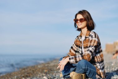Sahil kayalıklarında rahat bir kadın, gün ışığında okyanus manzarasına hayran, moda tarzı, doğa arka planı ve huzurlu ruh hali. Parlak gökyüzü, yumuşak rüzgar sakin.