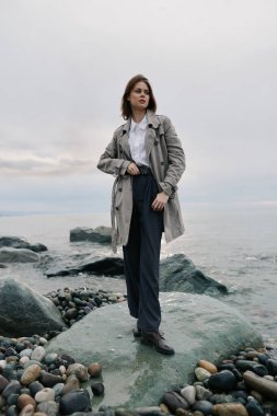 Koyu renk ceketli ve koyu renk pantolonlu genç bir kadın bulutlu gökyüzünün altında kayalık sahilde duruyor. Moda anlayışı, sonbahar stili, doğa ve barışçıl açık hava ortamı..
