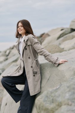 Tüylü bir trençkot ve siyah pantolon giyen mutlu genç kadın kayalık sahilde rahatlıyor. Bulutlu hava ve doğal arka plan gündelik tarz ve açık hava eğlence anını vurguluyor.