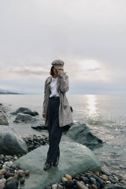 Uzun paltolu ve desenli şapkalı şık bir kadın arka planda deniz olan kayalık sahilde poz veriyor. Açık hava yaşam tarzı ve mevsimlik kıyafet konsepti için günlük sonbahar kıyafeti.