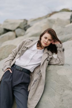 Sıradan genç bir kadın kayaların yanında duruyor. Palto ve beyaz gömlek giyip sakin bir ifade sergiliyor. Doğal ışığa sahip açık hava ortamları gevşek atmosfer ve yumuşak renkleri vurguluyor.