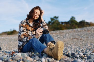 Dışarıdaki fotoğrafçı ve kadın, çakıl taşlı bir plajda, kamera, ekose gömlek ve botlar, doğal ışıkta sakin bir şekilde, sakince ışıldayan bir portre..