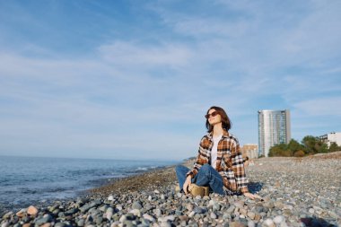 Dalgalar ve ufuk çizgisi olan kayalık bir kıyı şeridinde sıradan bir kadın sahnesi, katmanlı kıyafetler, meditasyon pozu, güneşli öğleden sonra, deniz manzarası, moda ve seyahat havası..