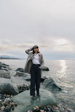 Şık paltolu ve şapkalı kadın okyanus kıyısında taşın üzerinde poz veriyor. Bulutlu gökyüzü ve yumuşak dalgalar, sakin havayı ve doğal manzaralı açık hava moda kavramını vurgular.