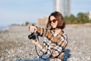 Güneşli bir sahilde kameralı, ekose gömlekli ve güneş gözlüklü bir kadın fotoğrafçının günlük açık hava portresi yaratıcılık, seyahat havası ve gün ışığında doğa.