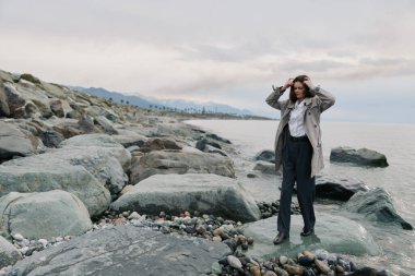 Yağmurluk ve koyu renk pantolon giyen genç kadın bulutlu gökyüzünün altında sakin deniz kıyısında duruyor. Moda, doğa, sonbahar, açık hava yaşam tarzı ve düşünceli ruh hali..