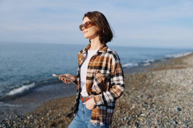 Sahil kenarında gezici bir cihaz, güneş gözlüğü, ekose gömlek ve kot giyen rahat bir kadın okyanus manzaralı ve güneşli bir gökyüzü olan açık hava anı.
