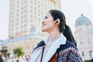 Bir kadının müzik dinlediği açık hava sahnesi kulaklıklar, şehir sokakları ve arkadaki ufuk çizgisi, şık palto, kendine güvenen bakış, rahat yaşam tarzı ve seyahat ilhamı.