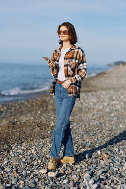Sahil sahnesinde telefon, güneş gözlüğü, ekose gömlek ve kot pantolon giyen bir kadın açık gökyüzünün altında ufuk ve deniz meltemiyle rüzgarlı bir kıyı şeridinde duruyor..