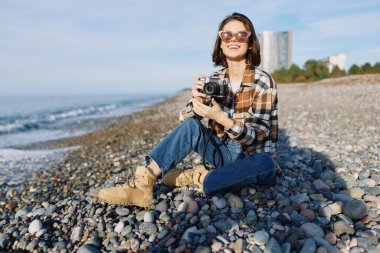 Kayalık bir sahilde fotoğrafçı bir kadının sıradan dış portresi, mercekleri hazır, güneşli deniz manzarasını yakalıyor ve tarzıyla ve enerjisiyle samimi anları yakalıyor. Doğal ışık şimdi dokuyu ve havayı geliştiriyor.