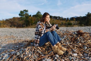 Kayalık bir sahilde kamera ile dinlenen kadın bir fotoğrafçı, ekose gömlek ve botlar giyiyor, açık hava çekimi sırasında kıyı dokularını ve ışığı belgeliyor.