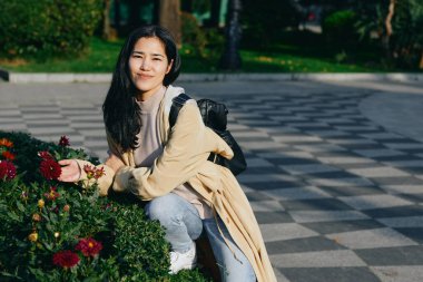 Parkside sahnesinde çiçeklerin yanında sırt çantalı genç bir kadın poz verirken hafifçe gülümsüyor, günlük kıyafet, açık hava manzarası, güneş ışığı, yeşillik, sakin atmosfer, günlük yaşam tarzı anı.