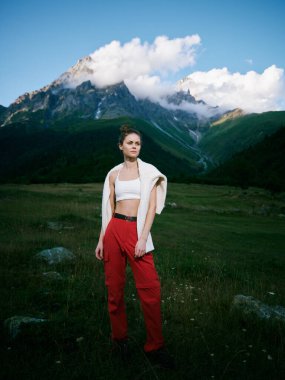 Açık hava moda sahnesinde kırmızı pantolonlu ve beyaz bluzlu bir kadın, arka planda dağlar, çayır, ışık, kendine güvenen bir tarz ve maceraperest bir ruh hali var..