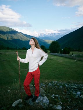 Beyaz süveter ve kırmızı pantolonlu bir kadınla açık hava keşfi sahnesi, bir vadi ve sarmal tepelerin yanında bir asayla dikilen, özgürlüğün altını çizen doğal manzara, yürüyüş ve