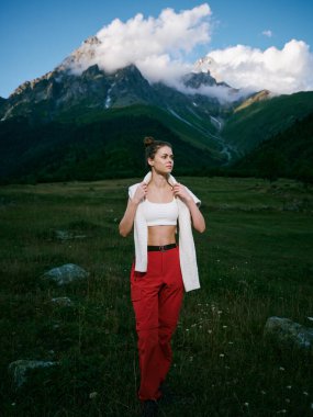 Manzaralı çayırlarda gündelik gezgin kırmızı pantolon ve beyaz bluz giyer, arka planda dağlar, geniş vadi ve temiz hava, doğa ve özgürlüğü yakalar..