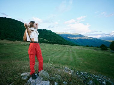 Kırmızı pantolonlu ve açık renk tişörtlü doğa yürüyüşçüsü, otlak bir ovanın üzerindeki uzak tepelere doğru bakıyor, yalnızlığı, merakı ve geniş vadide ve açık kırsalda doğayla bir bağı aktarıyor..