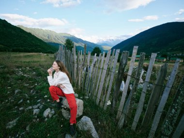 Dağların arasında bir vadide, ahşap bir çitin yanında bir kadınla açık hava manzarası, sakin bir ortam, ön planda kayalar ve çimenler, doğa yolculuğu, keşif ve hikaye anlatma için mükemmel bir manzara.