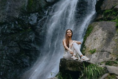 Genç bir kadın, arkasında çağlayan bir şelale ile sakince oturur, yemyeşil ve engebeli taşlarla kaplıdır, sükuneti ve doğayla bağlantıyı çağrıştırır.
