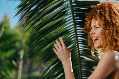 Kıvırcık saçlı gülümseyen kadın açık mavi gökyüzünün altında gür palmiye yapraklarının yanında duruyor. Güneşli tropikal ortamda neşe ve sıcaklık ifade ediyor. Doğal güzellik kavramı.