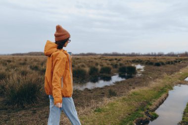 Young woman lifestyle scene outdoors walking near marshland wearing orange jacket and brown knitted hat. Casual autumn fashion and nature exploration create calm and reflective atmosphere.